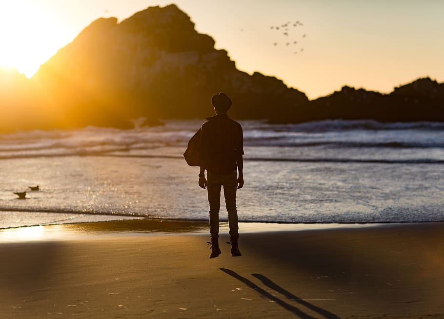 silhouette of man near seashore in front of mountain at daytime HD wallpaper