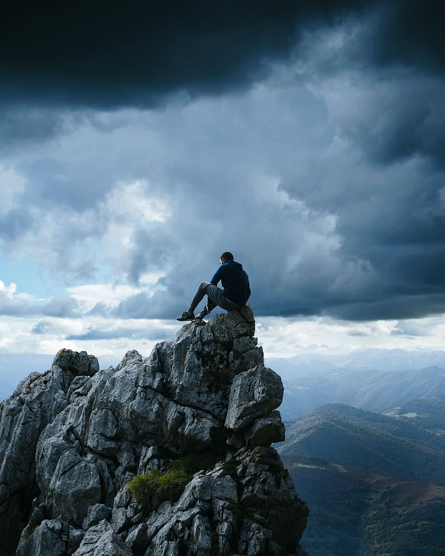 man wearing black pullover hoodie sitting on the high-top rock, man sitting on cliff under nimbus clouds HD wallpaper