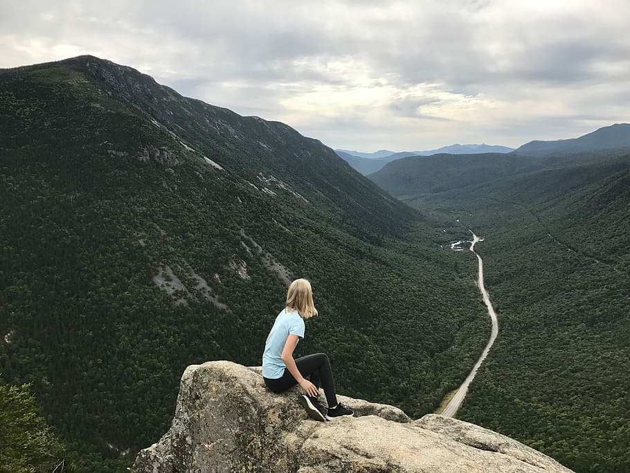 woman in blue t-shirt sitting on brown rock facing green mountains, woman sitting on cliff HD wallpaper