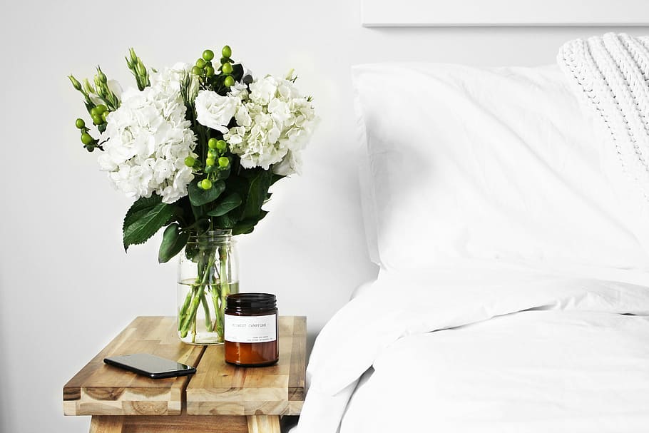 flowers in clear glass vase on table, white petaled flowers on vase on top of nightstand beside smartphone and bottle near to bed HD wallpaper