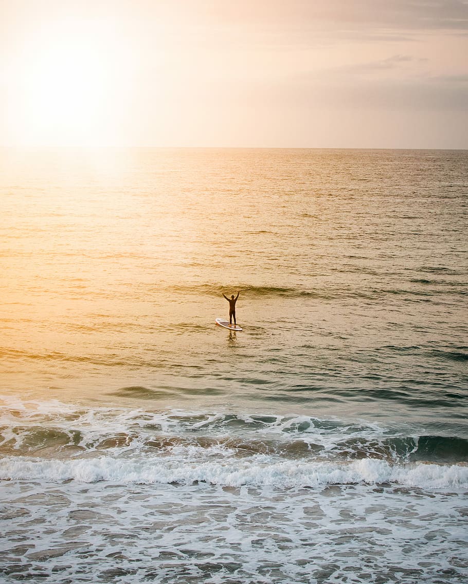 person standing on paddle board on body of water, man riding blue surfboard taken at sunset HD wallpaper