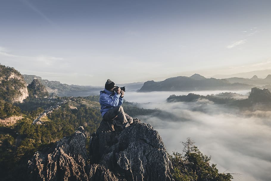 man on top of mountain taking pictures, man sitting on mountain peak holding camera