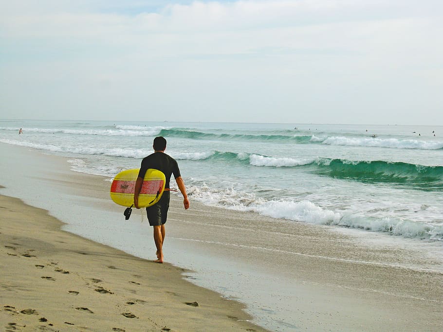 man carrying surfboard while walking on shore, man in black shirt and shorts carrying a yellow surfboard on beach HD wallpaper