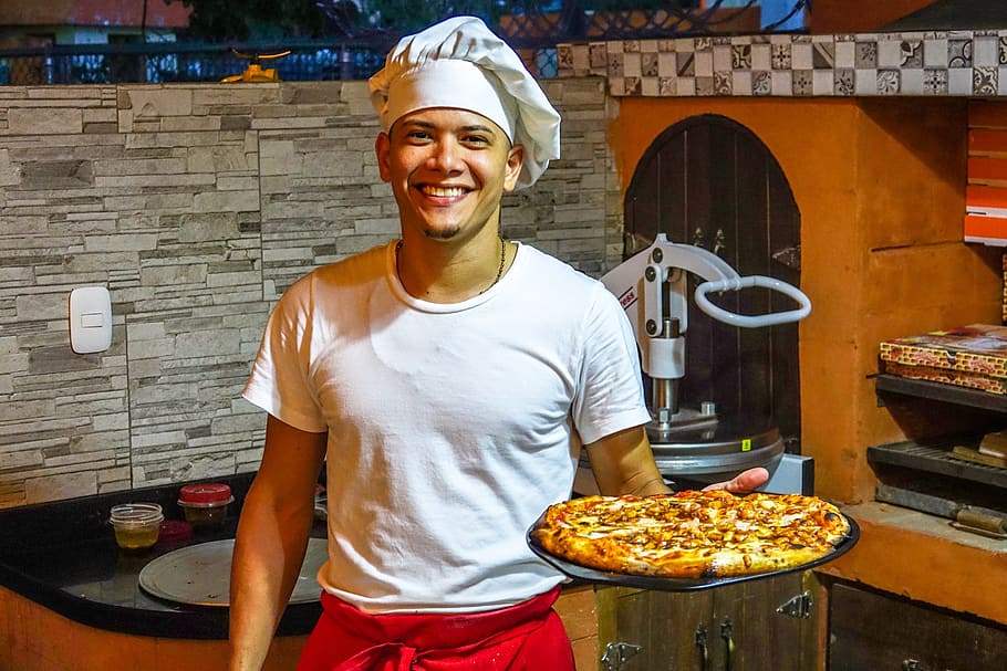 man holding baked pizza standing near black and brown wooden cabinet