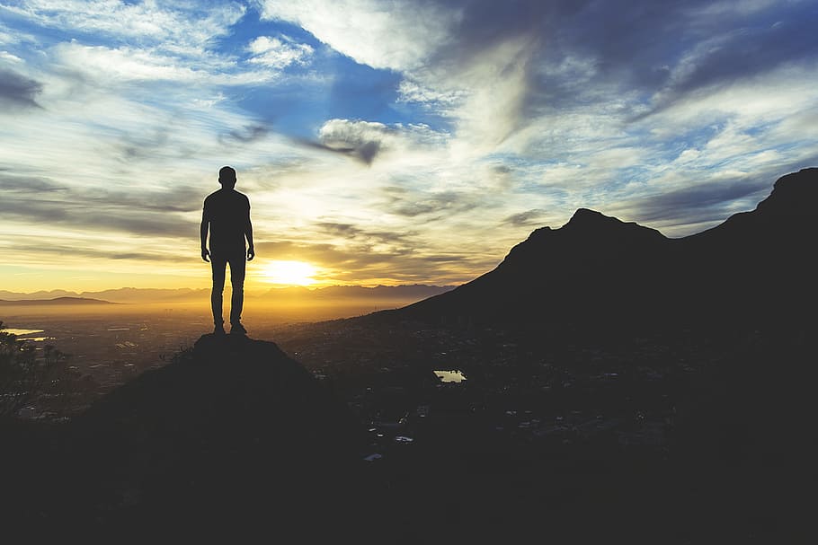 person standing at the top of mountain, silhouette photography of man standing on cliff during golden hour