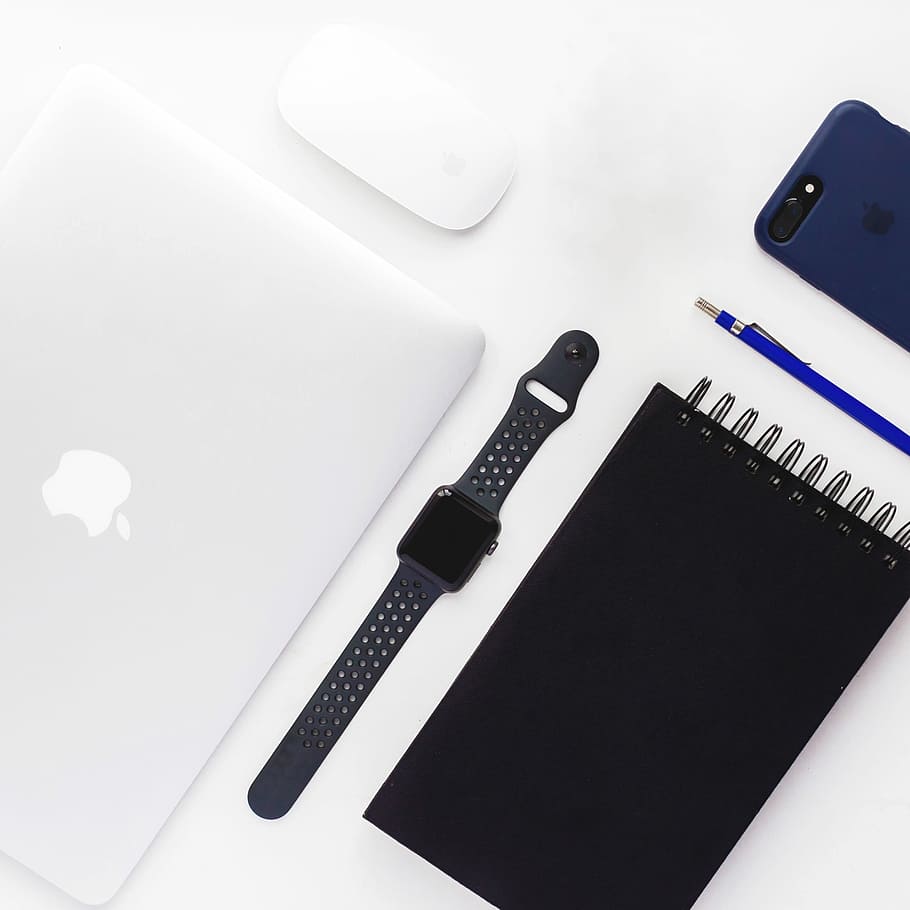 flat lay photography of space gray aluminum case Apple Watch with Nike Sport Band, spiral book, MacBook White, Apple Magic Mouse, and black iPhone 7 Plus with blue case, space gray Apple watch with black Nike+ Sport Band on top of white surface