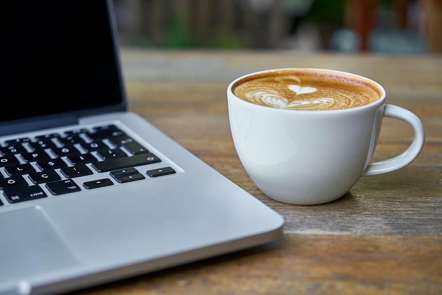 brown and white coffee in white ceramic mug beside of gray laptop computer HD wallpaper