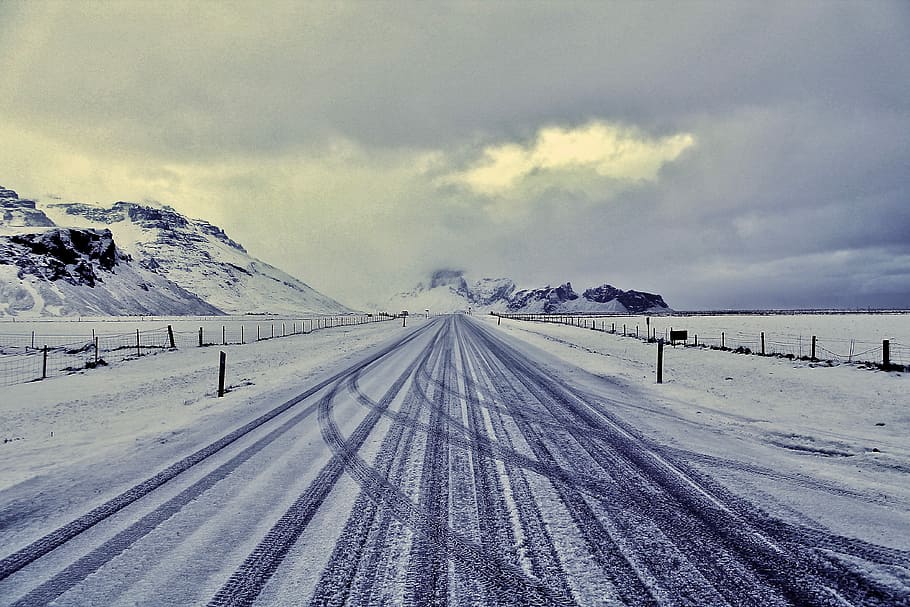 snow road with drift tire print near mountain range, snowcovered road under cloudy sky