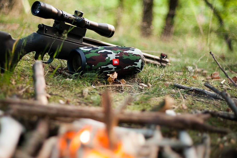 black sniper rifle beside green, black, and white camouflage JBL portable speaker, black rifle with scope beside JBL Charge 3 speaker
