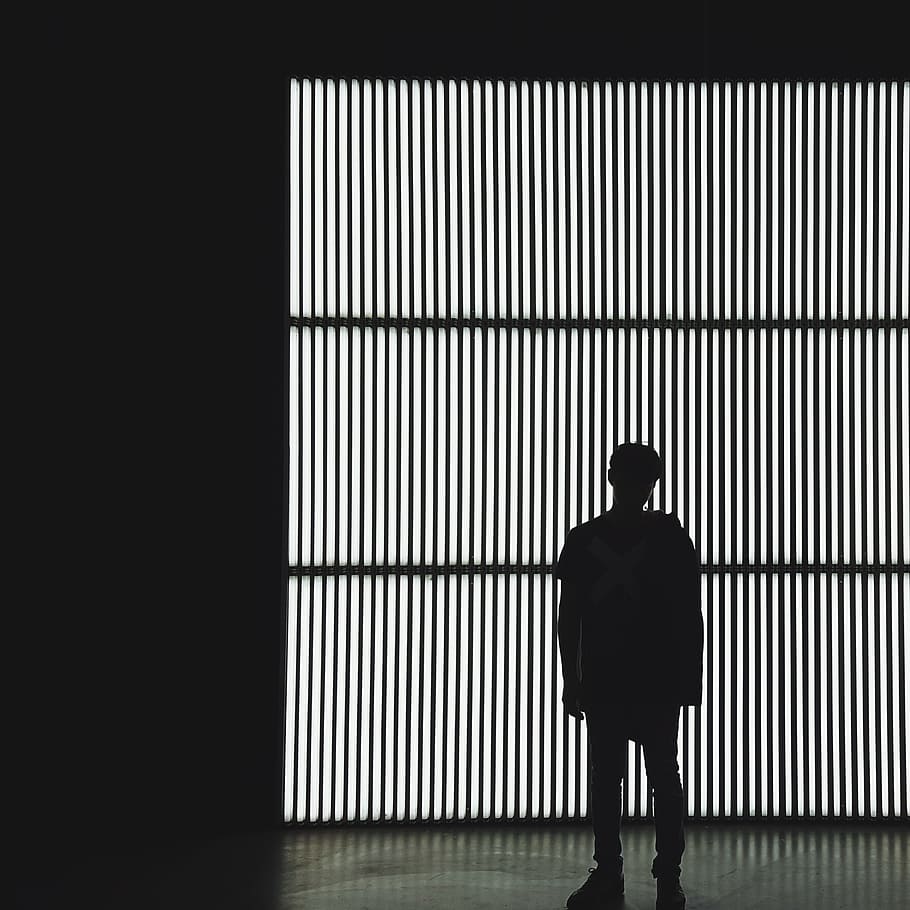 silhouette photo of a person standing near wall in dark room, silhouette man in jacket standing in front of light