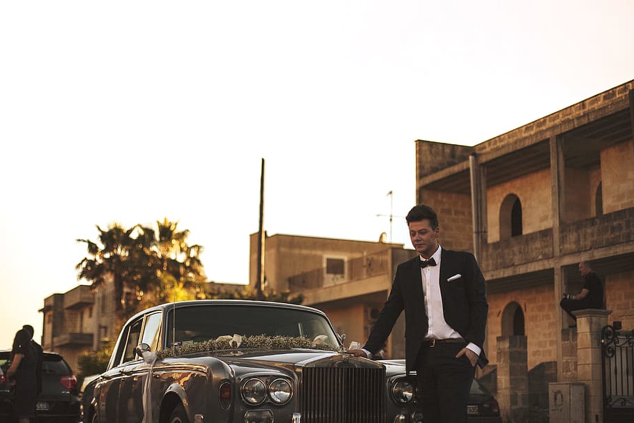 man wearing black suit jacket standing near gray car, man standing beside silver car HD wallpaper