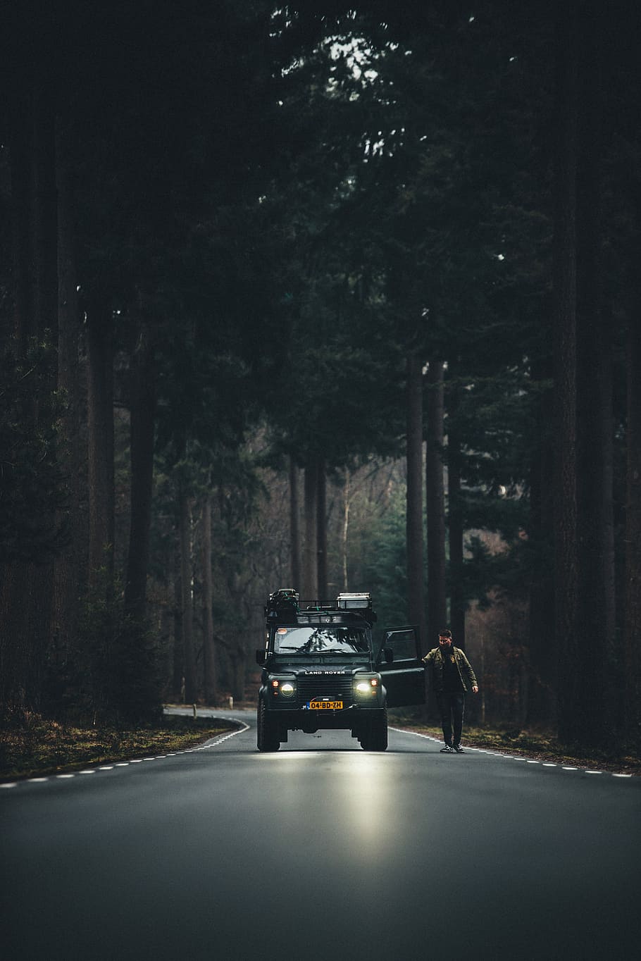 man in brown jacket standing beside car on road, man walking beside Land Rover SUV on road in between tall trees