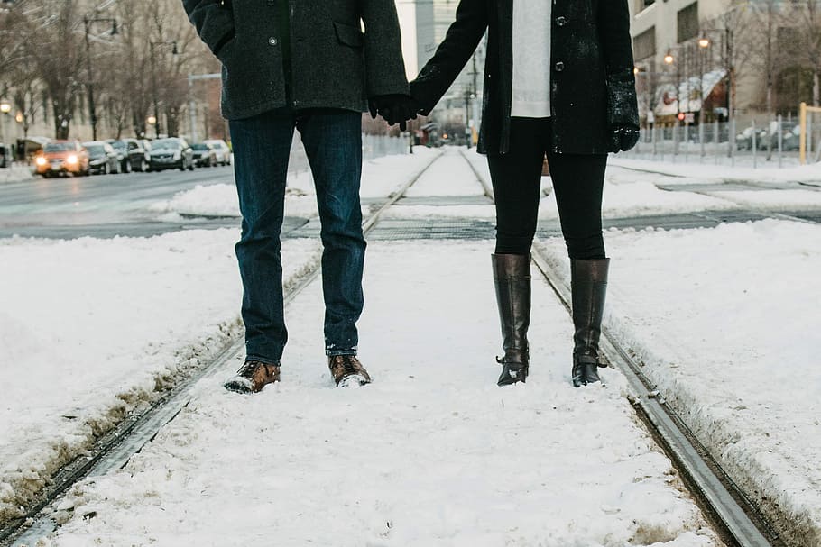 photo of two person standing on snow-covered road, couple holding hands while standing on snow field