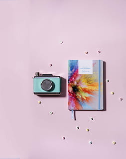 teal and silver-colored MILC camera beside paint powder burst book cover on pink surface HD wallpaper