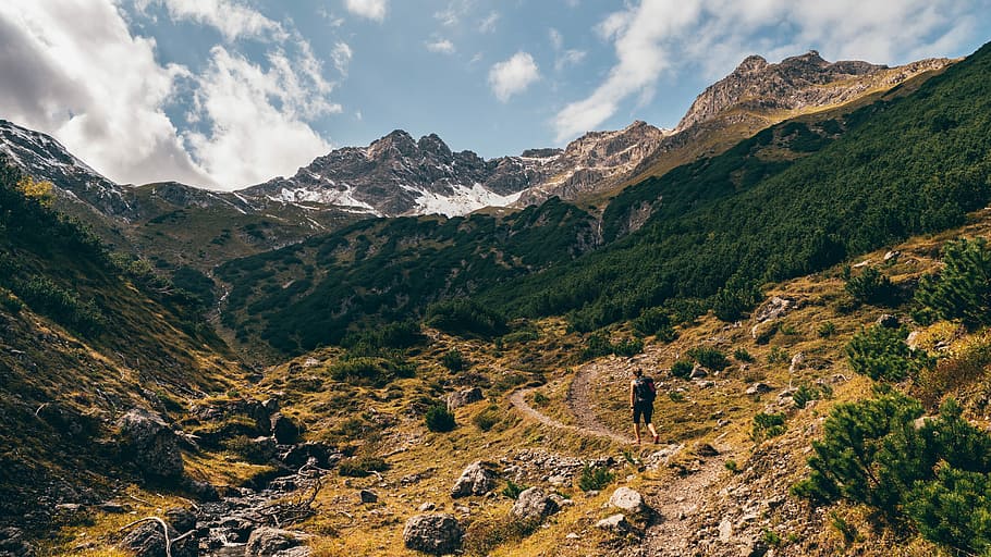man walking through dirt road near forest and mountain, man walking on hills near trees and grassed under cloudy sky during daytime HD wallpaper