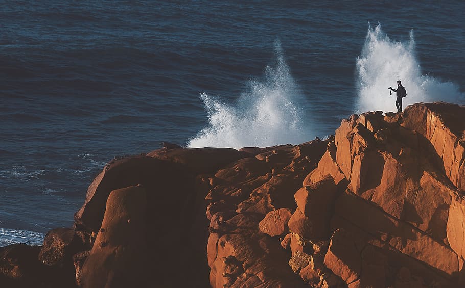 man standing on brown cliff near body of water during daytime, man standing on rocky mountain during daytime HD wallpaper