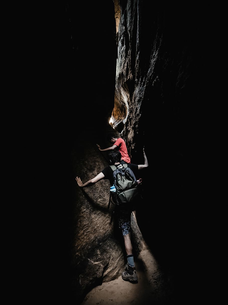 two boy's climbing between black and grey rocks during daytime, man in black shirt walking in the middle of the rock formation HD wallpaper