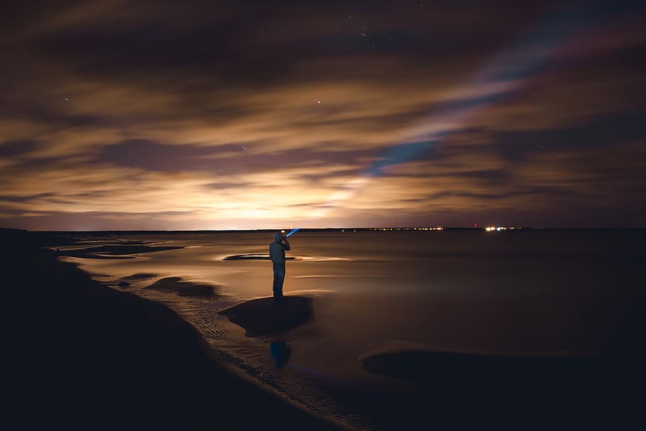 man standing on rock near body of water during golden hour, person standing on shoreline holding flashlight pointing upwards HD wallpaper