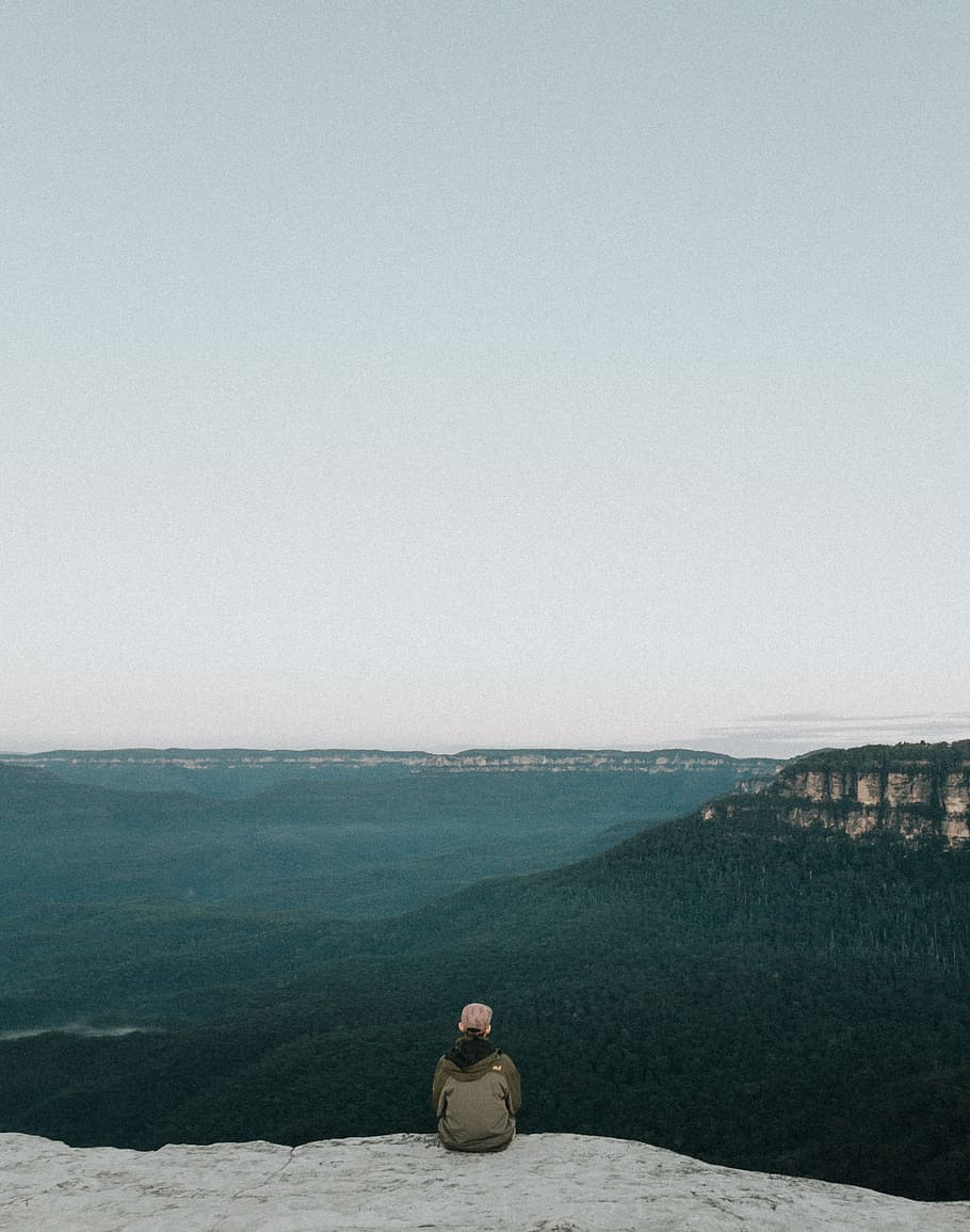 person sitting on rock while sight seeing, person wearing grey jacket sitting on edge of cliff facing mountains HD wallpaper