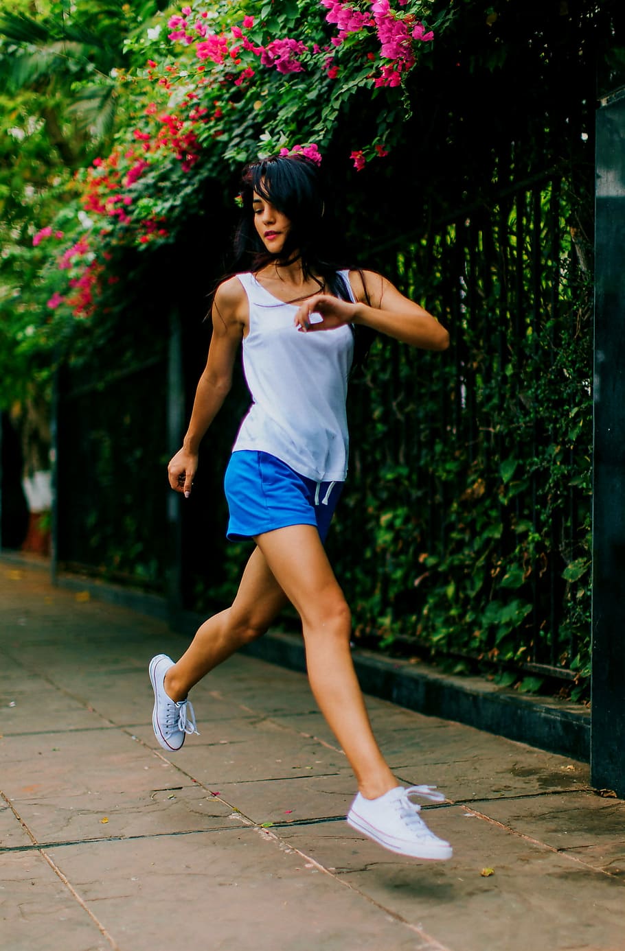 woman jumping on ground near black metal fence under pink petaled flower at daytime, woman wearing white tank top and blue short shorts outdoors HD wallpaper
