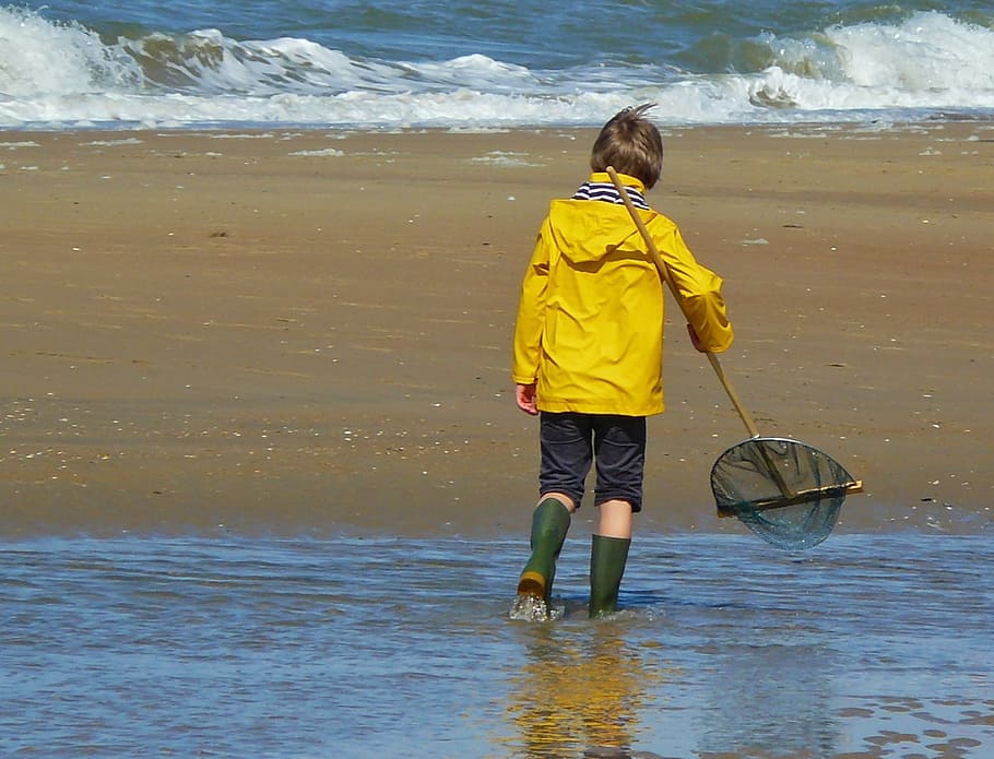 boy wearing yellow rain coat and blue capri pants with pair of green rain boots on body of water holding net