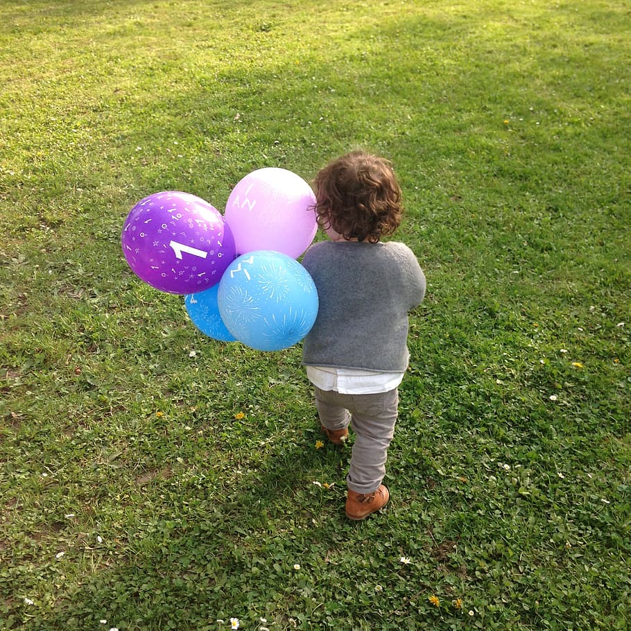 child holding assorted-colorful balloons while walking on green grass field during daytime HD wallpaper