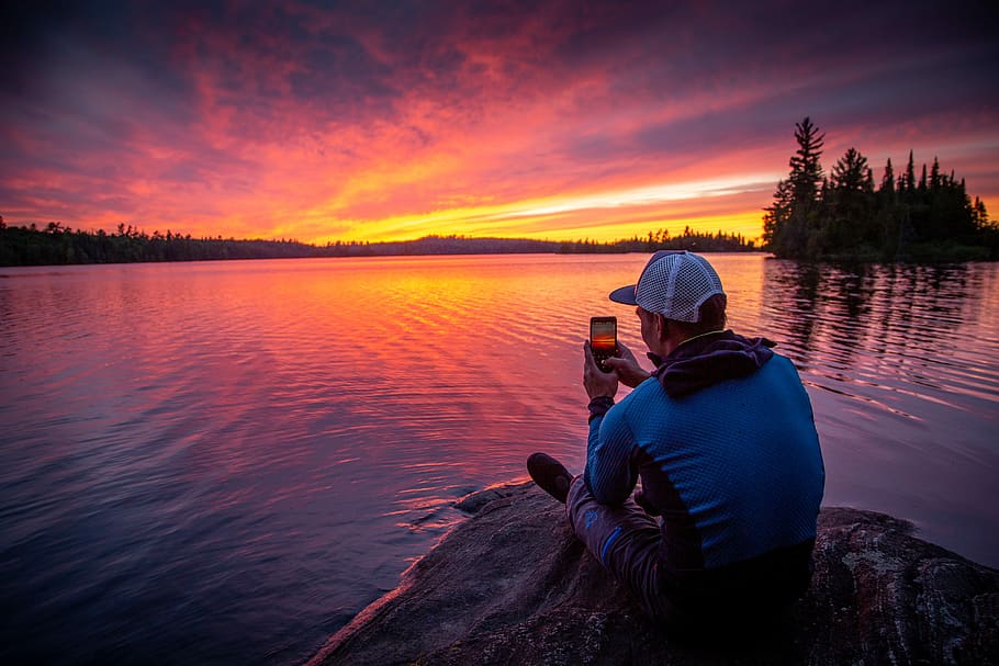 man sitting on gray rock while taking photo of silhouette, man sitting on rock formation front of calm water during golden hour HD wallpaper