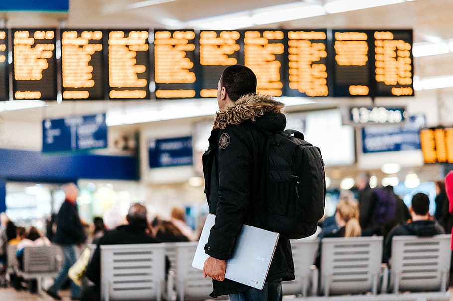 man standing inside airport looking at LED flight schedule bulletin board, man wearing black and brown parka jacket standing while looking at display monitor