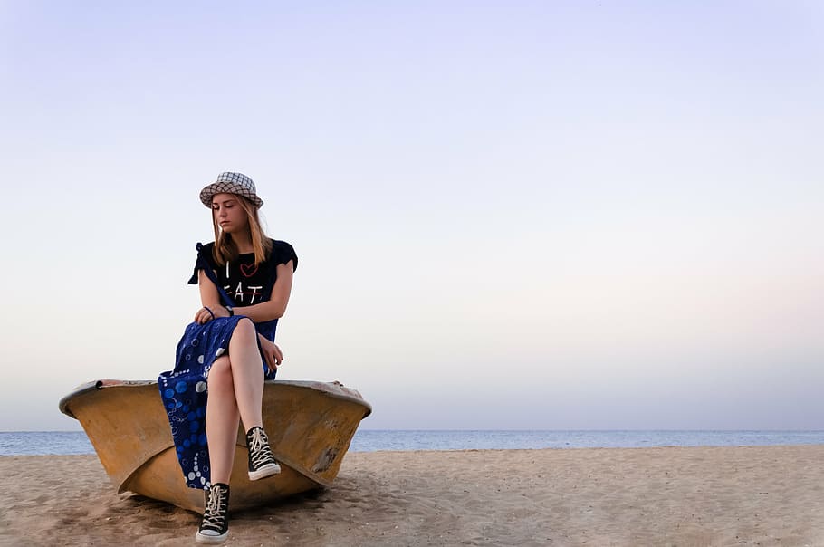 woman in black crew-neck T-shirt and blue skirt sitting on brown boat at the seashore under gray skies during day time HD wallpaper