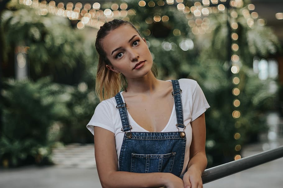 woman wearing bib and brace overalls leaning her face to left, selective focus photography of woman in denim overall leaning on metal hand rail