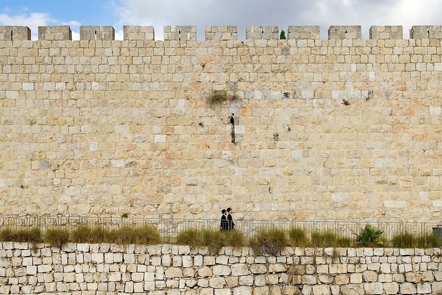 two person walking beside beige brick wall, two people walking near beige stone wall under gray clouds during daytime HD wallpaper
