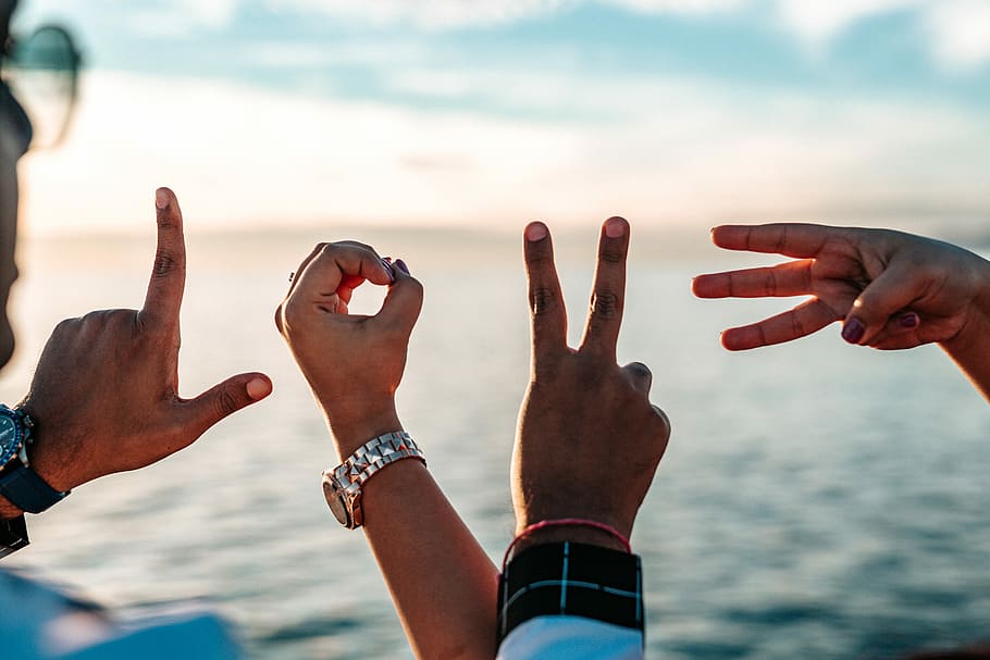 two persons forming love fingers, person with watch facing body of water doing hand sign