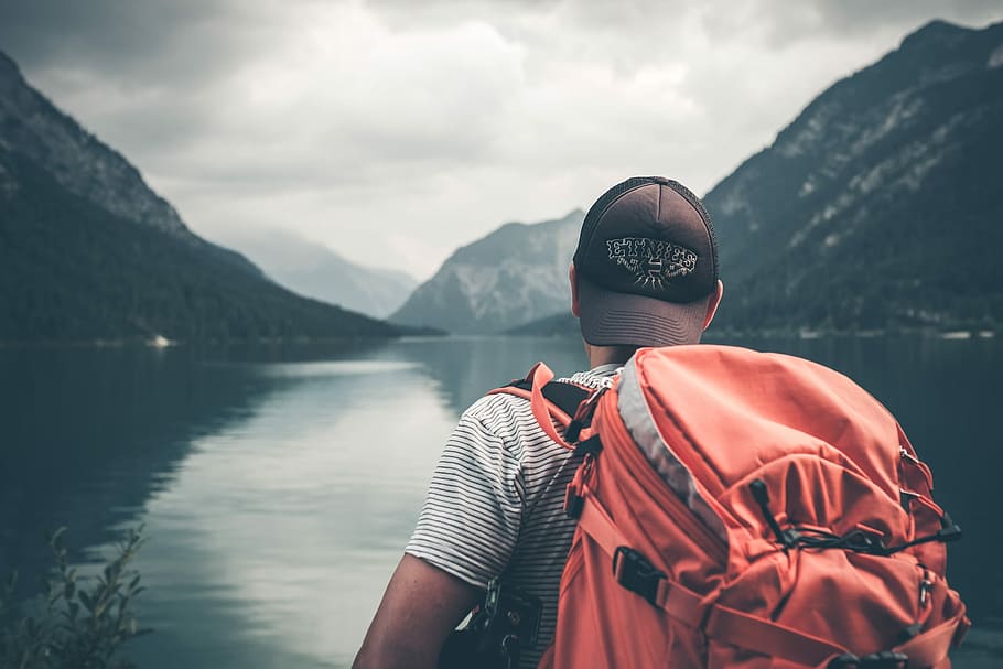 man with red hiking backpack facing body of water and mountains at daytime, man wearing orange backpack near body of water