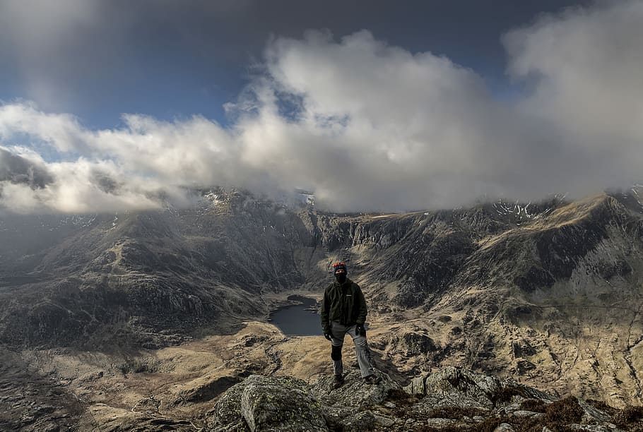 person standing on rocky mountain during daytime, man standing on rocky mountain overlooking lake during daytime HD wallpaper