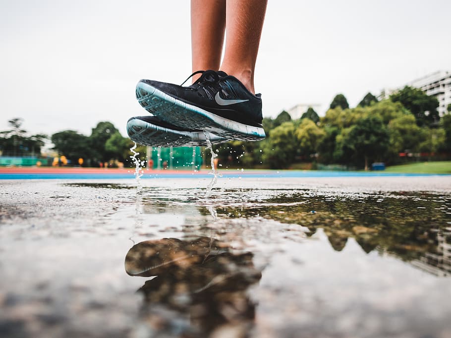 person wearing pair of gray-and-white Nike running shoes jumping on gray concrete floor with water during daytime, reflection photography of a person wearing sneakers