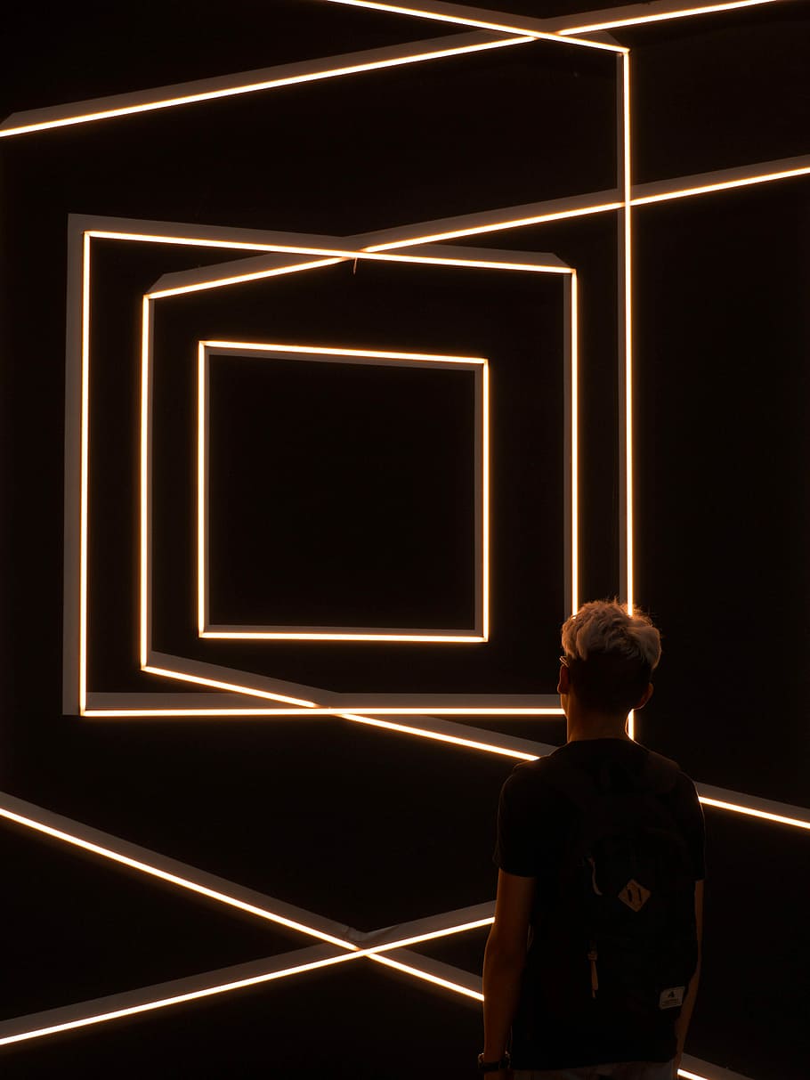 person standing in front of optical illusion wall, man in black top wearing backpack in dark room lowlight photography