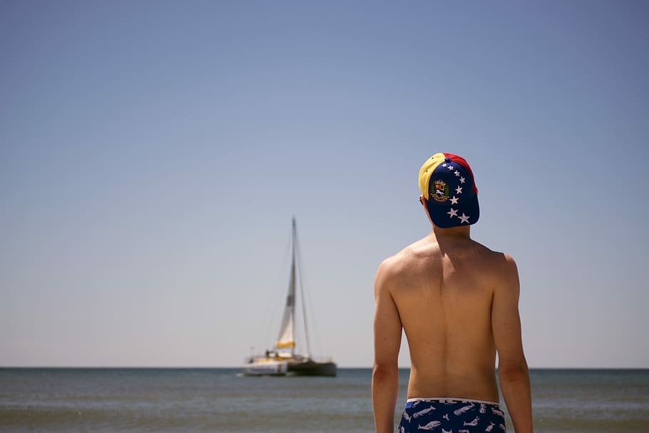 man standing facing boat during daytime, man with yellow, blue, and red cap standing viewing sail boat HD wallpaper