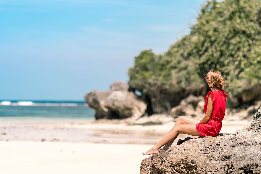 selective focus photo of woman sitting on rock formation seashore, woman in red rompers sitting on rock looking at sea