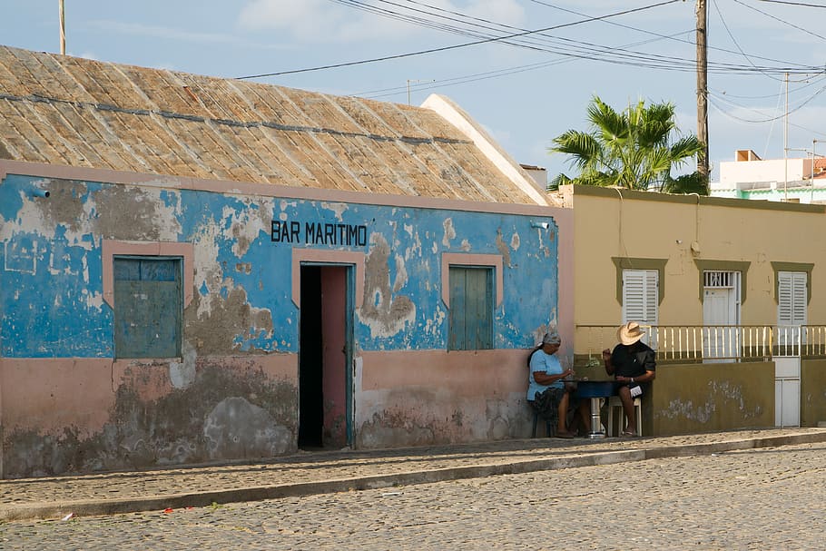 man and woman sitting outside house beside road, two person sitting beside brown concrete building during daytime HD wallpaper
