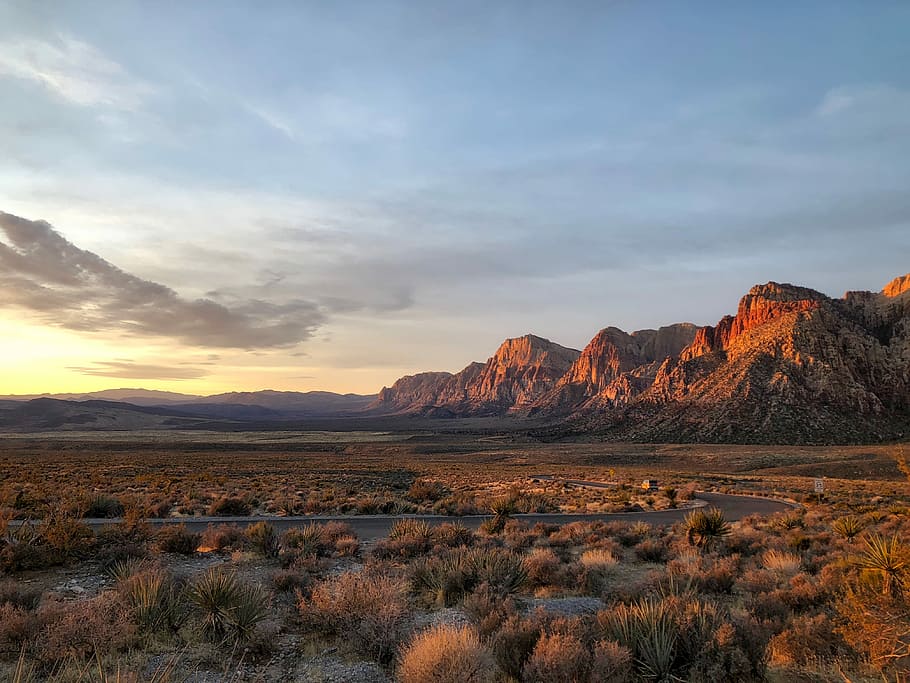brown rocky mountain under cloudy sky, gray road surrounded by beige and green fields overlooking rock formation during daytime HD wallpaper