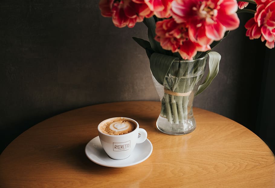 clear glass vase with red petaled flowers near cup filled with coffee both on round brown wooden table, cappuccino served on white ceramic cup HD wallpaper