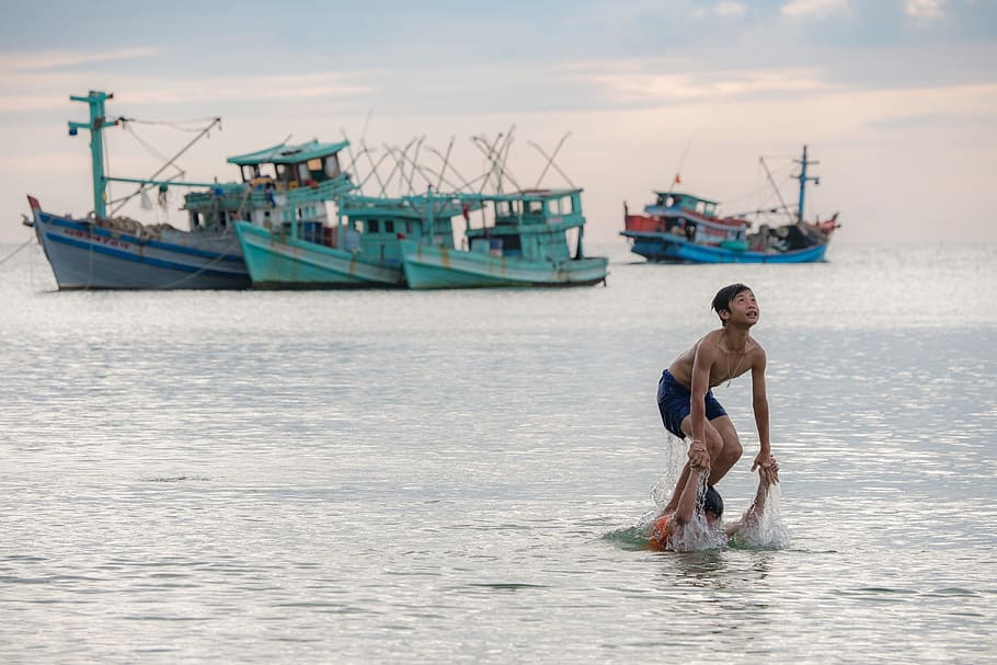 person swimming on sea near ships during daytime, boy standing on another person's shoulder at sea near boats at daytime HD wallpaper