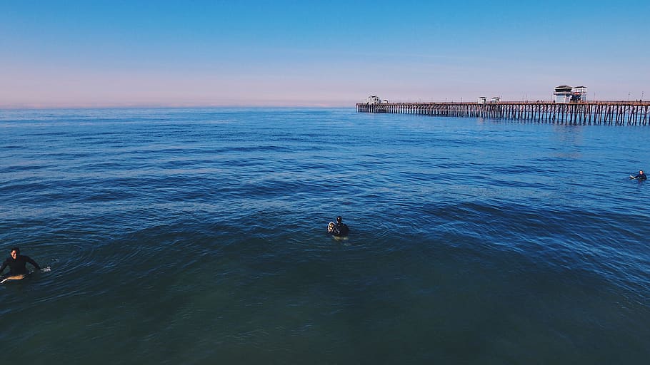 three people swimming on ocean under blue sky, three person on body of water near sea dock HD wallpaper