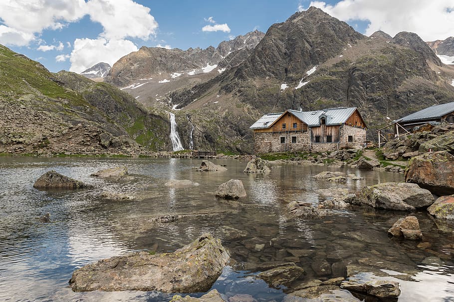 brown house near at body of water and surrounded by rocks during daytime, brown concrete house near river and mountains at daytime HD wallpaper