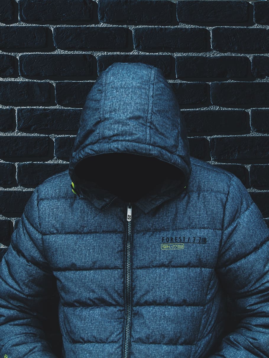 person with head down wearing blue zip-up jacket leaning on wall, man leaning on black concrete brick