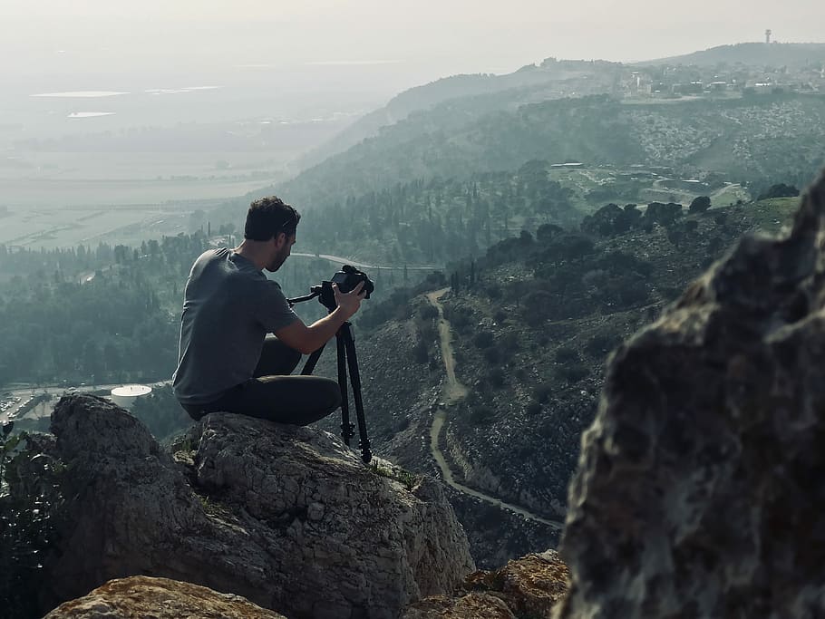 man sitting on rock using camera, man sitting on gray rock holding black DSLR camera with tripod HD wallpaper