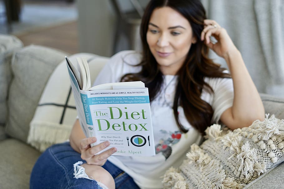 woman sitting on couch reading book, woman reading The Diet Detox book sitting on gray sofa HD wallpaper