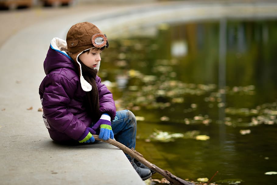 boy sitting on pavement while holding stick in front of body of water