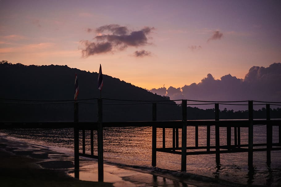 silhouette of wooden dock near mountain beside body of water during sunset, two multicolored flags on dock HD wallpaper