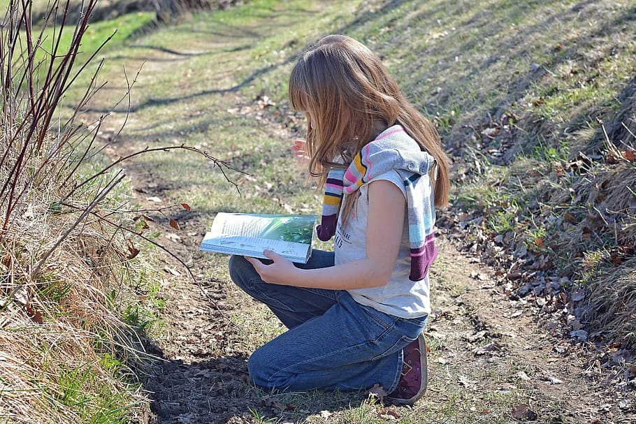 girl wearing white and brown stripe shirt and blue denim jeans reading a book while sitting in the ground during daytime
